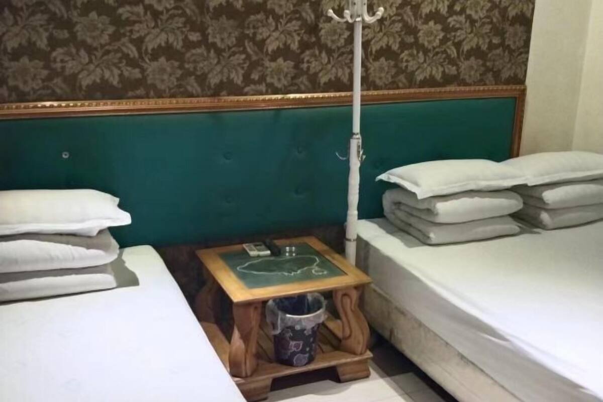Une chambre à coucher avec une tête de lit recouverte d’un tissu vert, une table de chevet avec une lampe et des serviettes blanches soigneusement pliées.