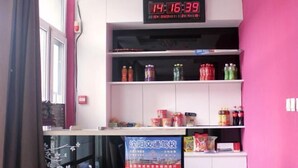 Ein Zimmer mit einer digitalen Uhr, die 14:16:39 anzeigt, Regalen voller Getränke und Snacks sowie einem Poster an der Wand.