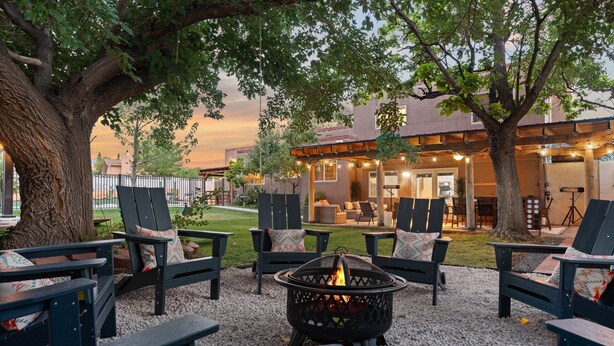 A cozy outdoor seating area with Adirondack chairs and a fire pit, surrounded by trees and a building with a covered patio. - Luxury Amenity Paradise! Sleeps 18 - Pool, Hot Tub, Game Room, Golf, Fire Pit!
 (Albuquerque)