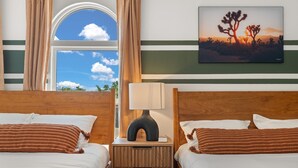 A hotel room with two beds, a nightstand with a lamp, a painting of a desert landscape, and a window with curtains. - Luxury Amenity Paradise! Sleeps 18 - Pool, Hot Tub, Game Room, Golf, Fire Pit!
 (Albuquerque)