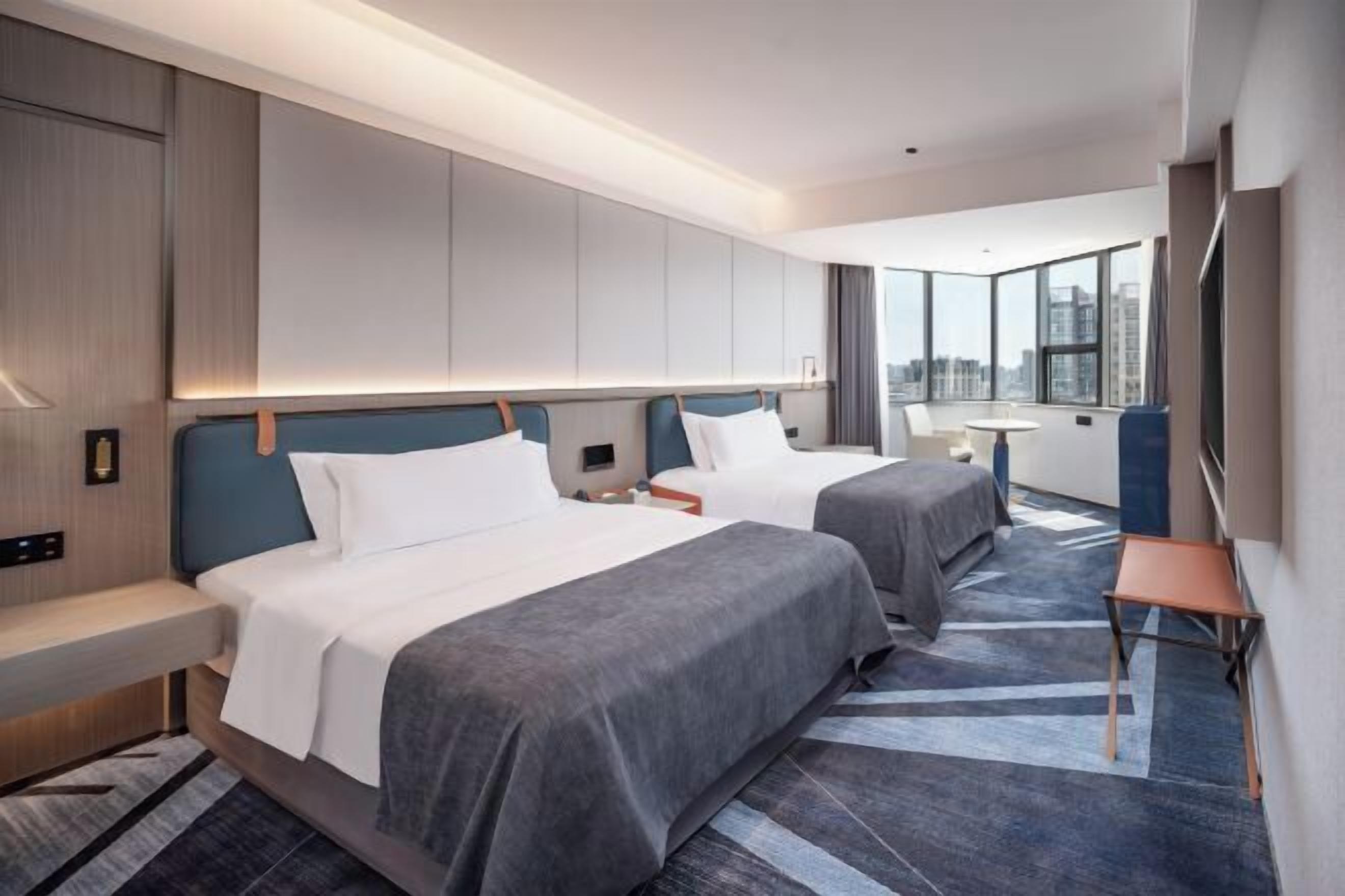 Ett modernt hotellrum med två sängar, ett skrivbord och ett stort fönster med utsikt över staden.