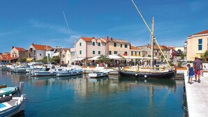 Un port de plaisance où sont amarrés plusieurs bateaux, une jetée où se promènent des gens, et des bâtiments aux toits rouges.