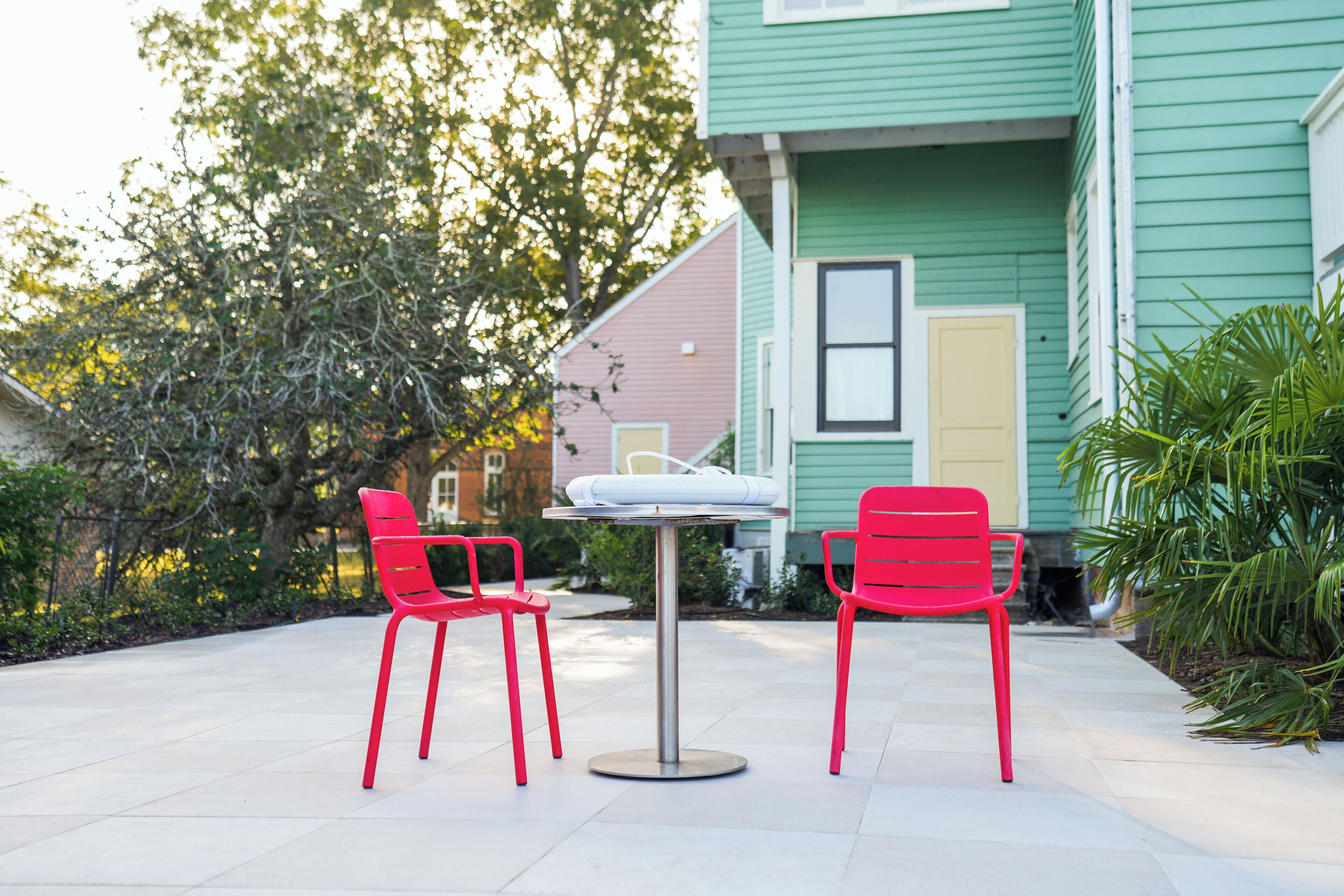 An outdoor patio with two red chairs and a table, surrounded by greenery and a house with green siding.