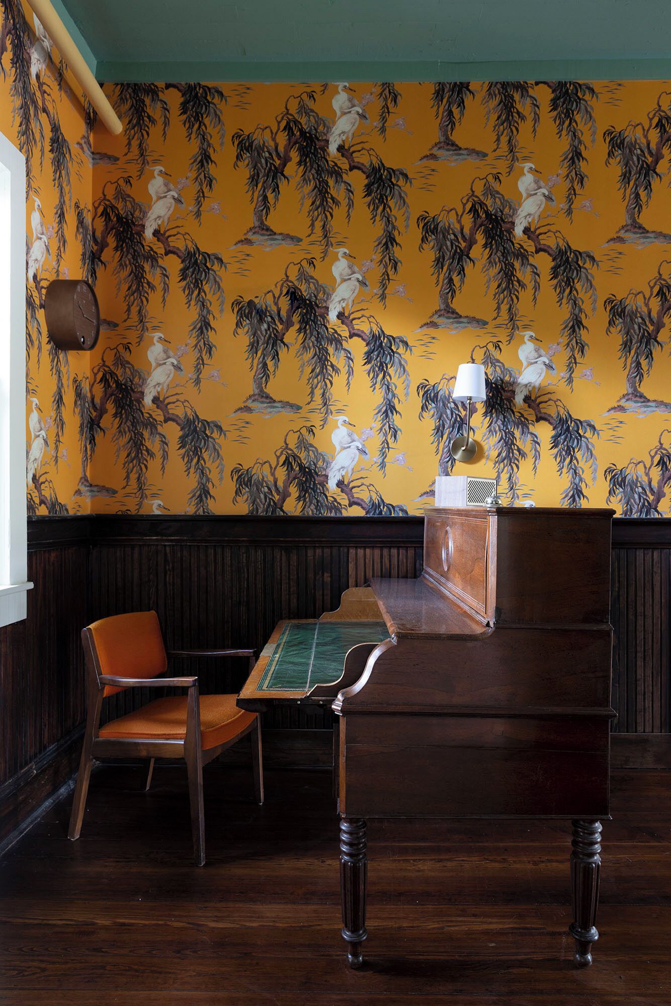 Quarto com um piano de madeira, uma poltrona verde estofada e uma parede com papel de parede floral amarelo.