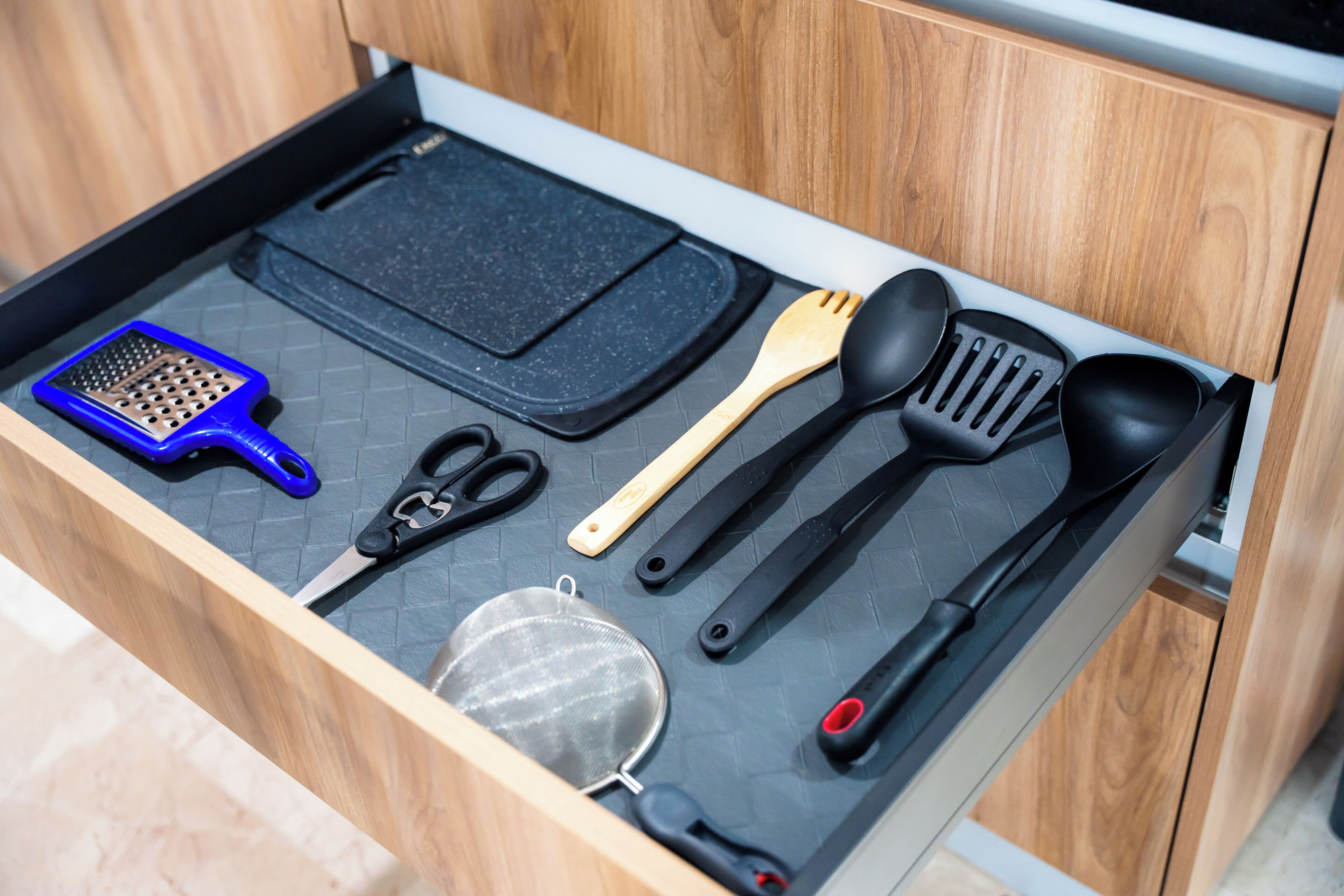 Un cajón de cocina con un tapete negro, un rallador, tijeras, un colador, una espátula de madera y una cuchara para servir negra, junto con otra espátula negra.