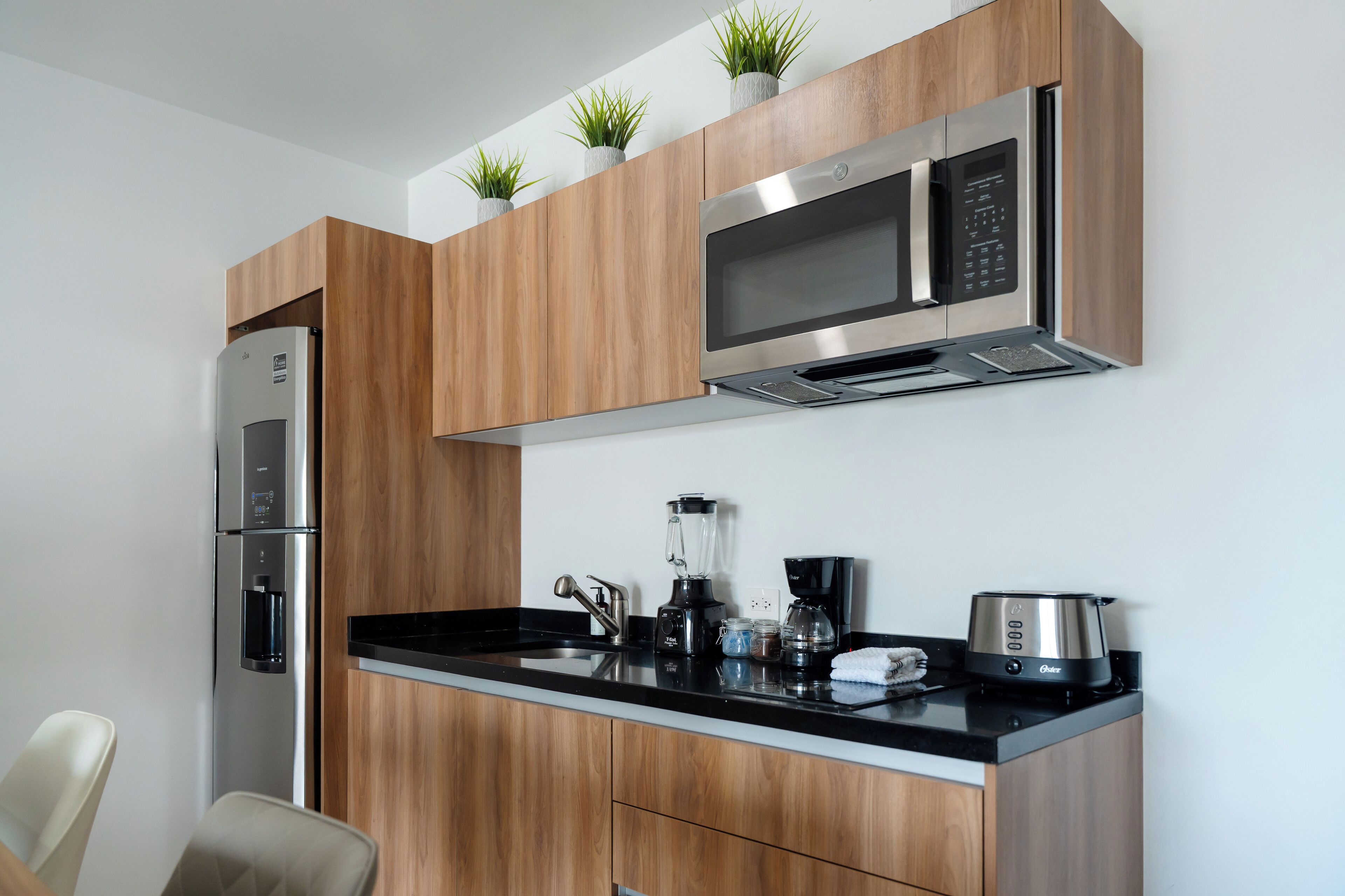 A modern kitchen with wooden cabinets, a stainless steel refrigerator, a microwave, a coffee maker, and a blender.