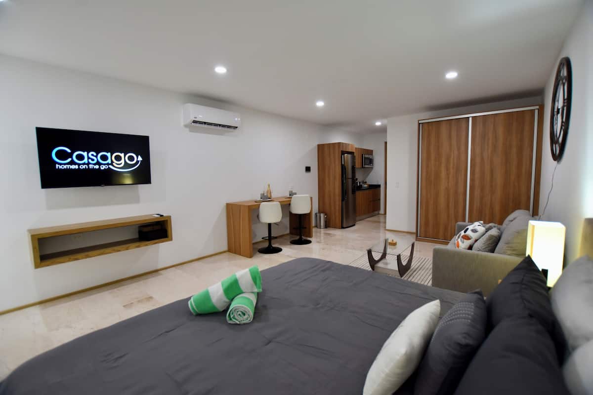 A modern hotel room with a large bed, a TV mounted on the wall, a wooden wardrobe, a small desk with a chair, and a sofa.