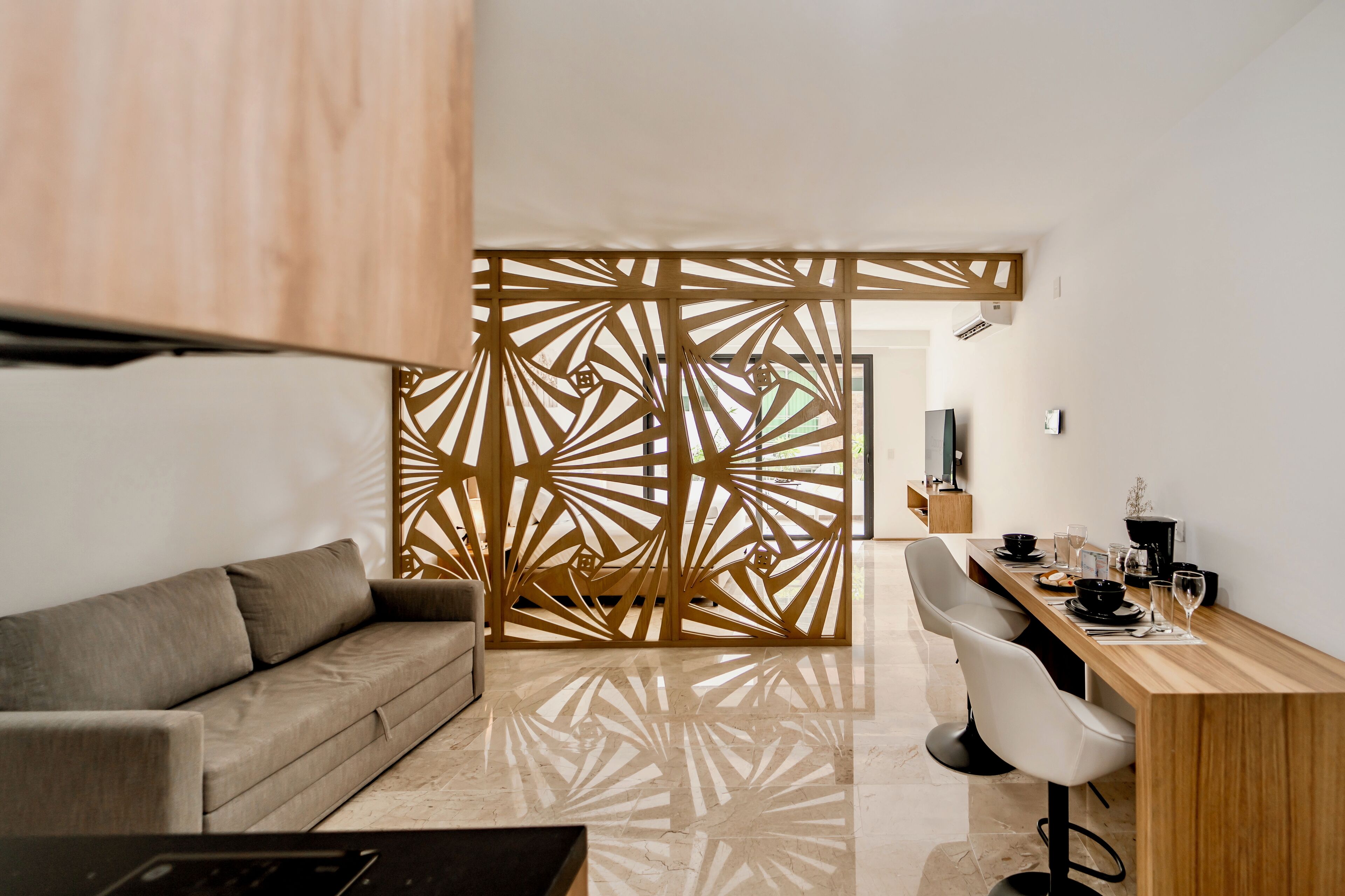 Una sala de estar moderna con un sofá, un separador de madera con diseños geométricos, una mesa de comedor con sillas y un televisor en la pared.