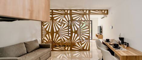 Una sala de estar moderna con un sofá, un separador de madera con diseños geométricos, una mesa de comedor con sillas y un televisor en la pared.
