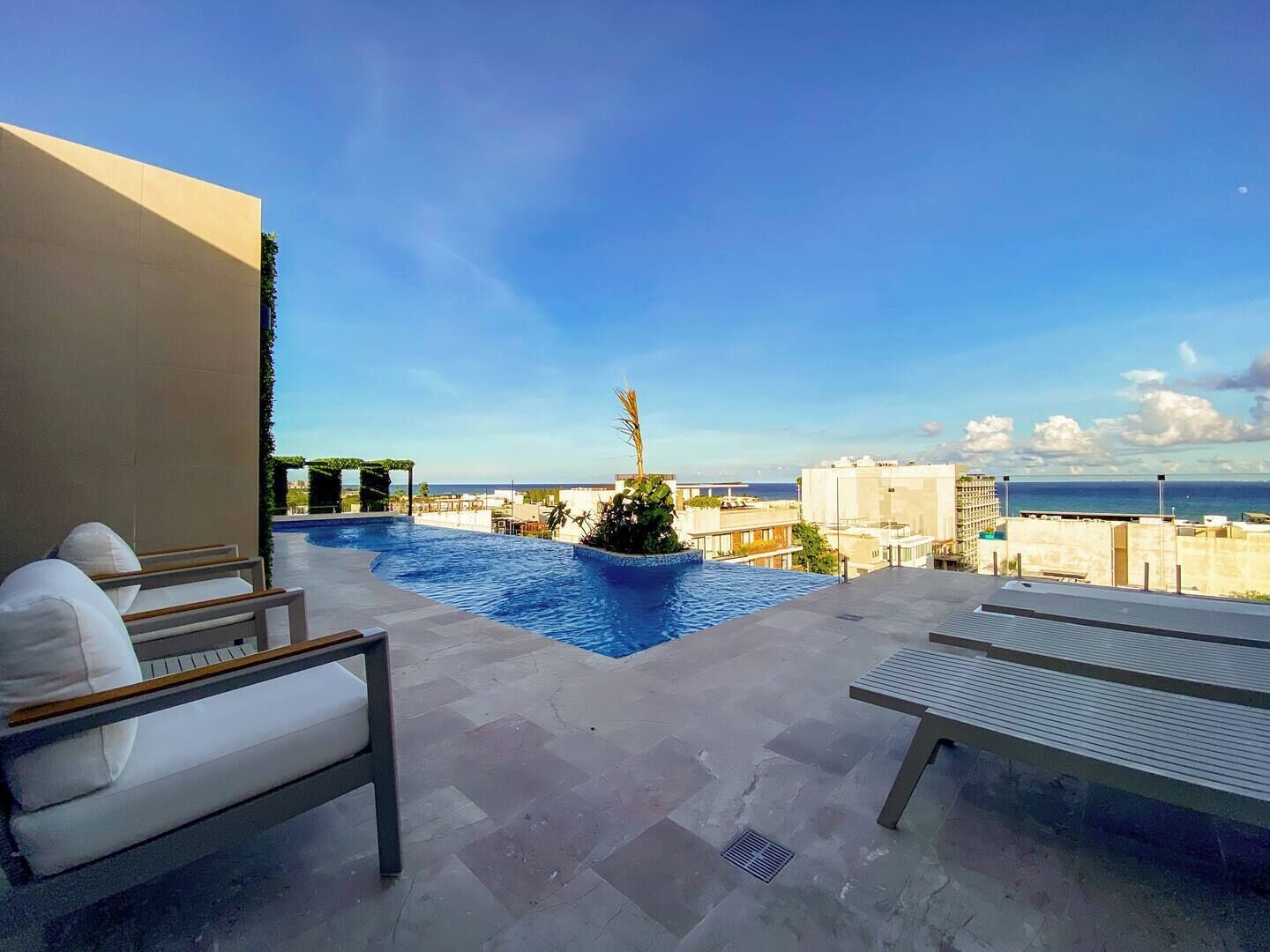 A rooftop pool area with lounge chairs, a small water feature, and a view of the city and sea.