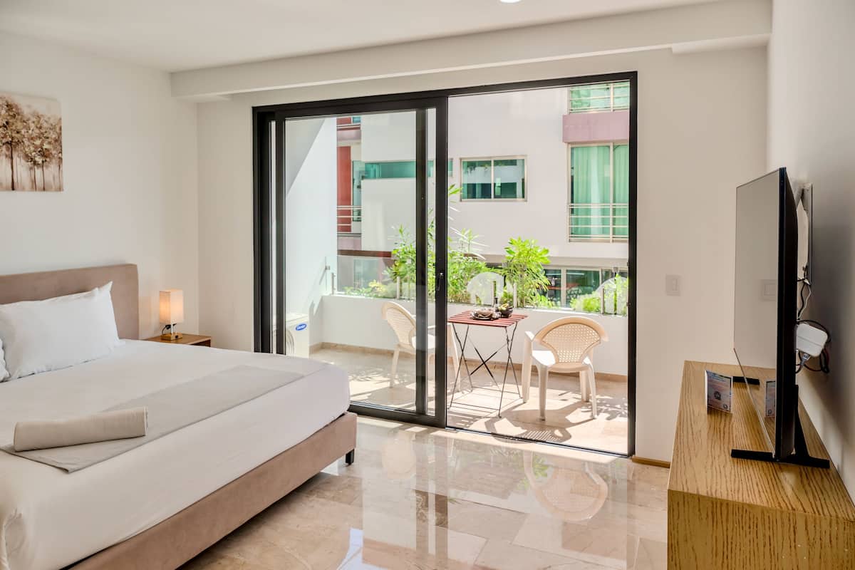 A modern hotel room with a large bed, a flat-screen TV, a balcony with a table and chairs, and a view of the building outside.