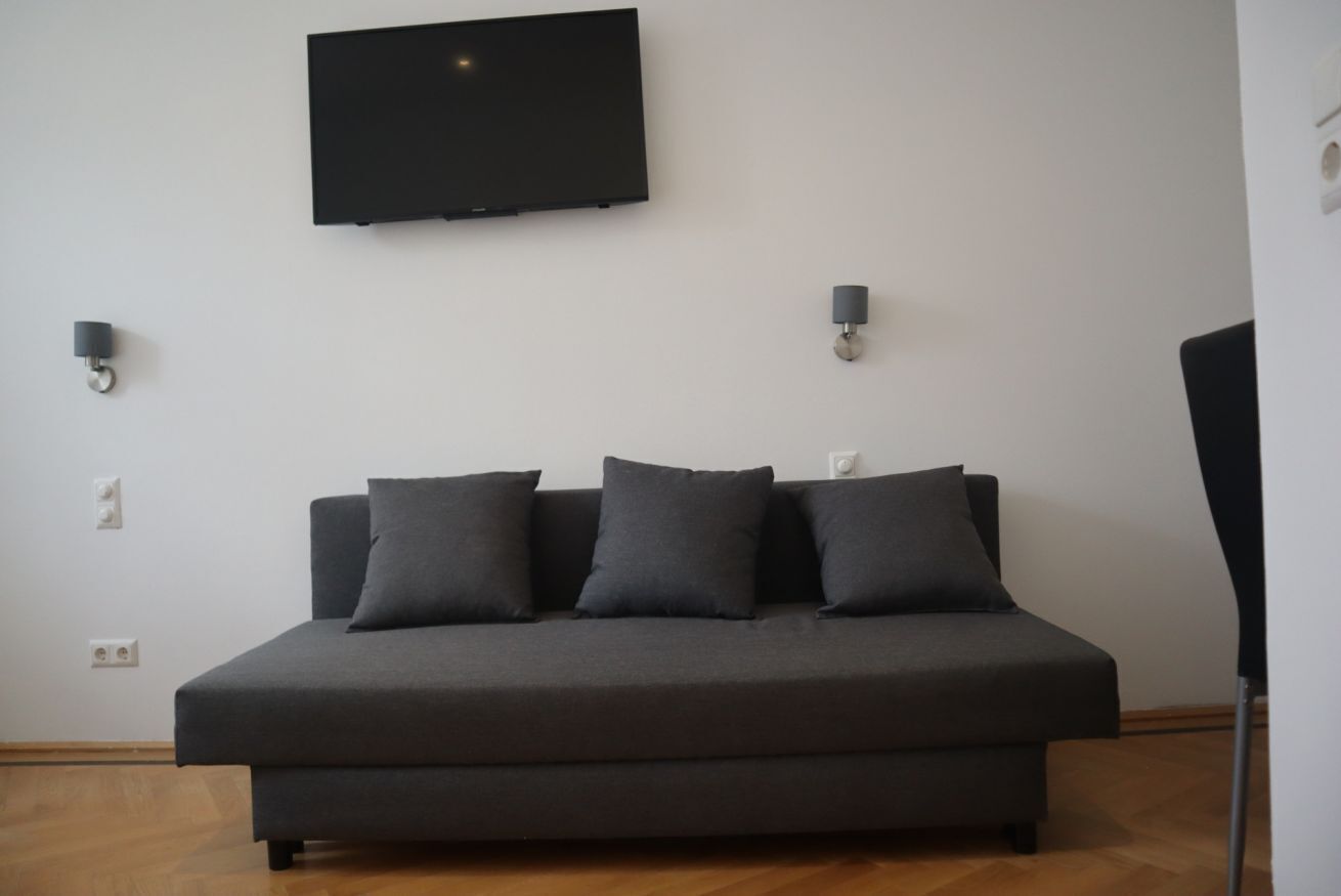 A modern room with a dark sofa, a flat-screen TV mounted on the wall, and two wall-mounted lamps.