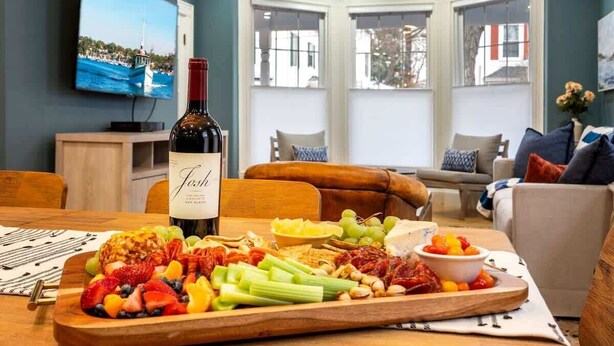 A dining area with a wooden table set with a variety of fruits and cheeses, a bottle of wine, and a television displaying a scenic view. - Workspace | Soaking Tub | W/D | 5 Min Downtown (Portland)