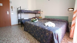 Una habitación con literas, una cama con colcha estampada, una mesita con un jarrón de flores y una puerta.