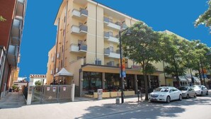 Un hotel de varios pisos con balcones, una entrada visible y coches aparcados en la calle.