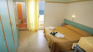 Un dormitorio con cama, mesita de noche, ventana con cortinas, puerta que da al baño y vista al mar.