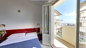 Una habitación de hotel con una cama, una mesita de noche, una lámpara de pared y vistas al balcón y edificios exteriores.