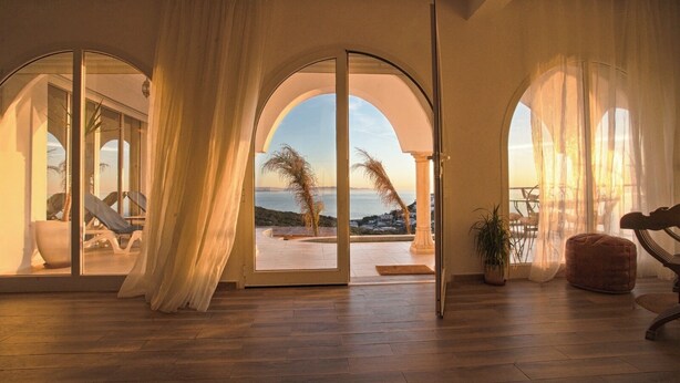 A room with wooden flooring, large arched windows with sheer curtains, a view of the sea, and outdoor seating with a palm tree. - Magnifique Villa Avec Piscine et vue sur mer & Montagnes (Béja Governorate)
