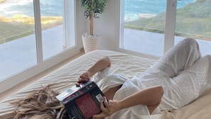 A person lying on a bed with a book, reading, in a room with large windows overlooking a coastal view. - Magnifique Villa Avec Piscine et vue sur mer & Montagnes (Béja Governorate)