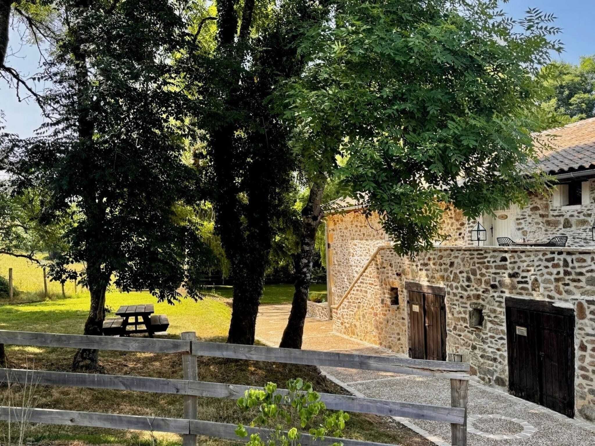 Un bâtiment en pierre à l’aspect rustique, avec une clôture en bois et une table de pique-nique, le tout situé dans un cadre verdoyant et luxuriant.