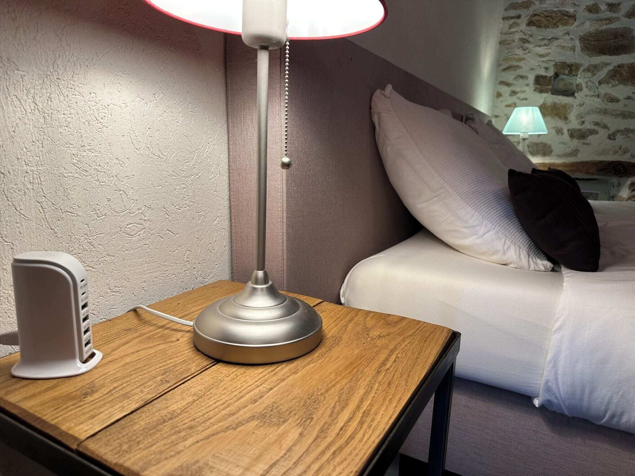 A wooden bedside table with a lamp, a white bedside phone, and a bed with white and black pillows.
