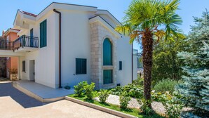 Una casa moderna de dos pisos con balcón, fachada de piedra y un jardín bien cuidado con diversas plantas y una palmera.