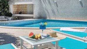 Un área junto a la piscina con una mesa dispuesta con una bebida, sandía y piña, y una silla de descanso con una toalla.