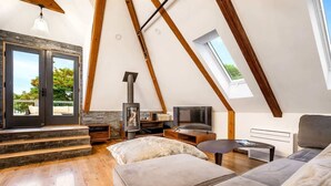 A loft-style room with a high ceiling, wooden beams, a fireplace, a television, and a large window.