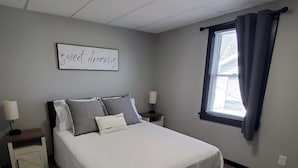 A bedroom with a bed, bedside tables, a wall-mounted light fixture, a window with curtains, and a wall sign. - Charming building with 1 bedroom and AC, WiFi in serene Whitehall (Whitehall)