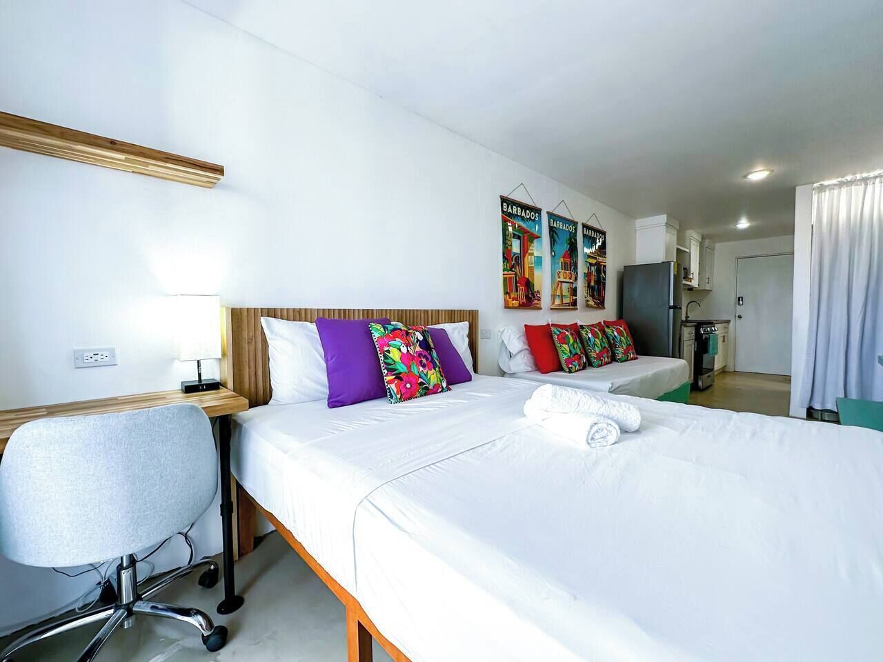 Habitación de hotel con dos camas, un escritorio y una silla. Hay almohadas coloridas y un cartel en la pared.