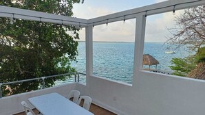 A balcony with a table and chairs, overlooking a body of water and a thatched structure. - ZENSUS LAGUNA (Bacalar)
