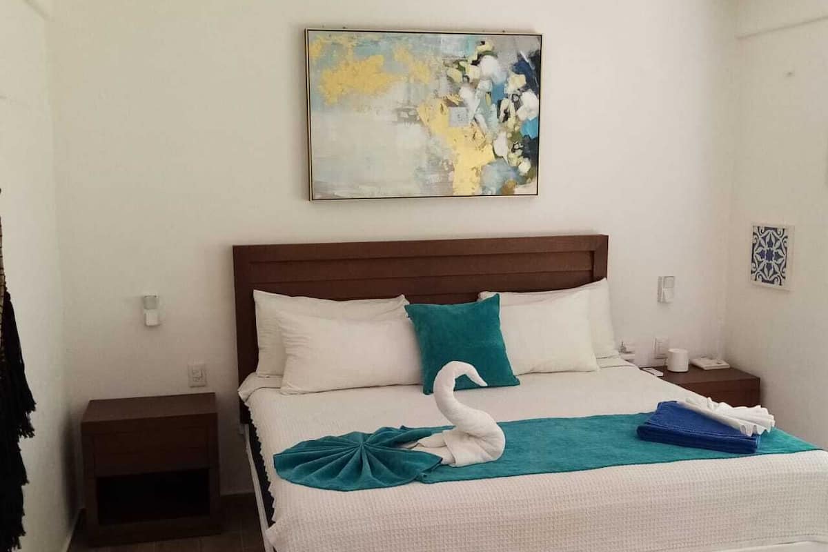 A neatly made bed with a swan-shaped towel and a teal pillow, a small bedside table, a wall-mounted abstract painting, and a patterned rug.