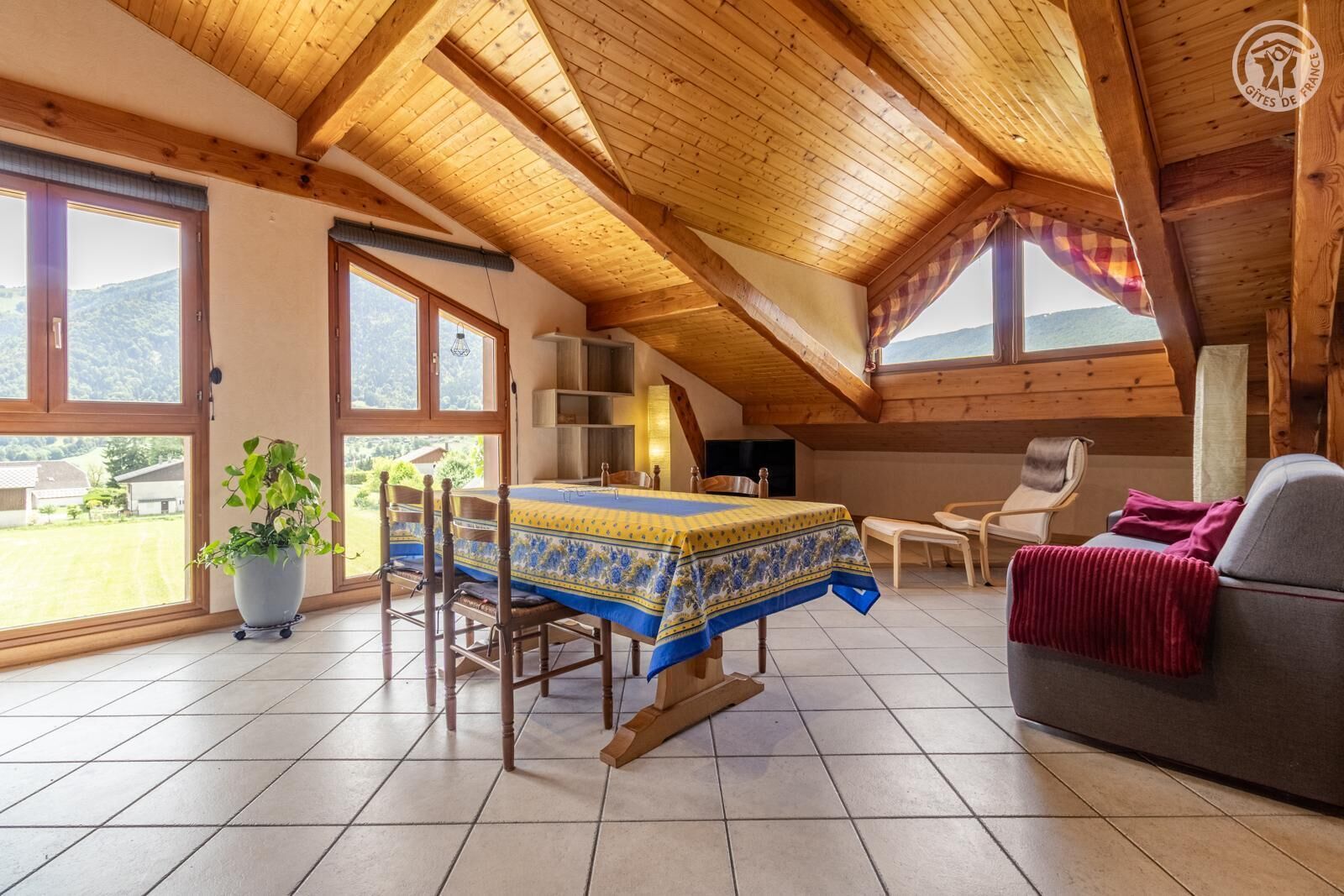 A cozy attic room with a wooden ceiling, a dining table with chairs, a sofa, and a large window offering a view of the outdoors.