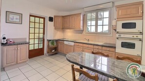 Een keuken met houten kastjes, een dubbele oven, een spoelbak en een eettafel met stoelen.