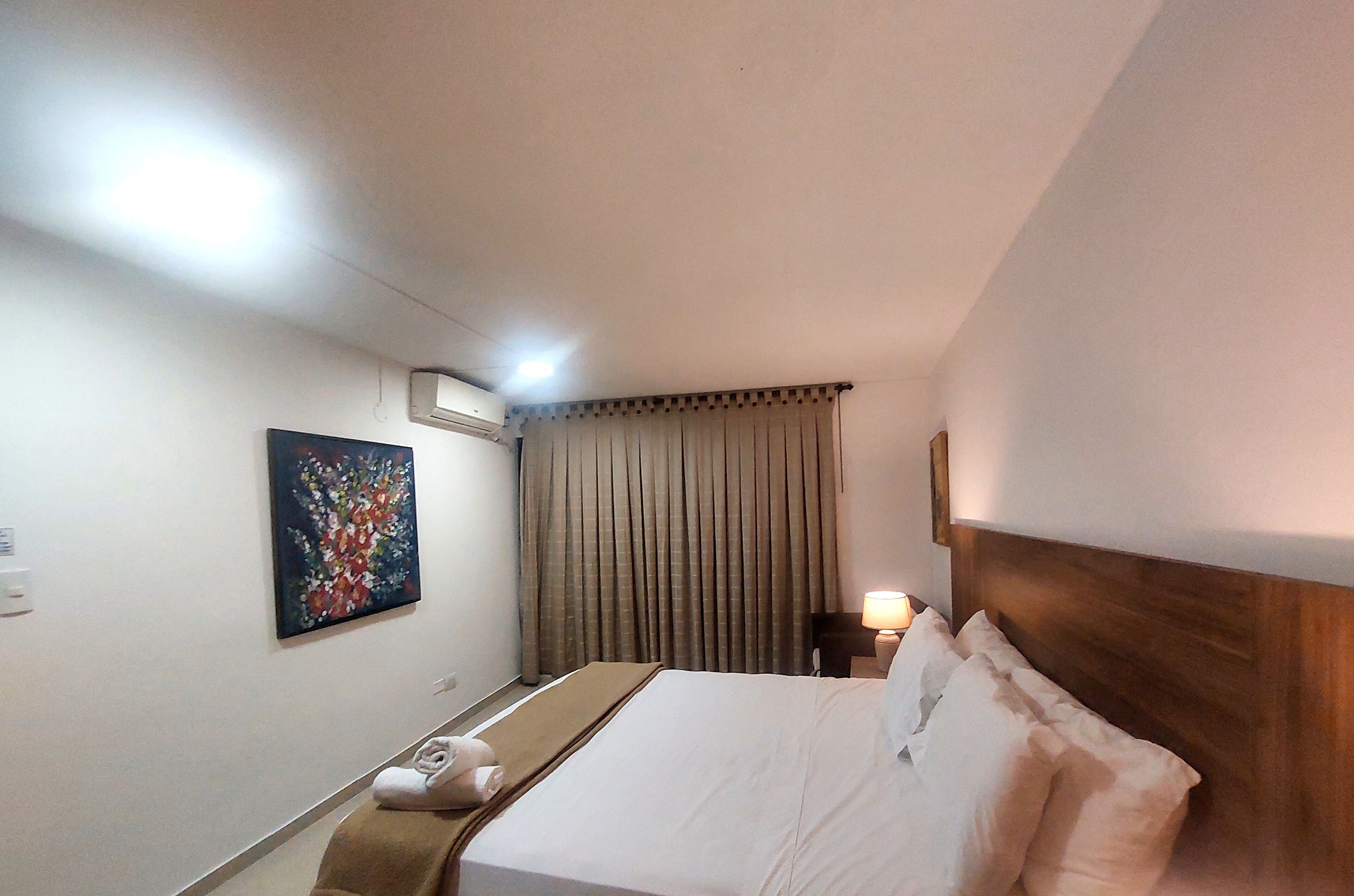 A hotel room with a bed, a nightstand, a lamp, a painting, and a window with curtains.
