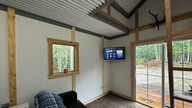A cozy room with a wooden ceiling, a window with a view of trees, a flat-screen TV mounted on the wall, and a black leather sofa with a blanket.