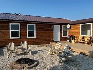 Una cabaña de madera con techo metálico, dos ventanas, una puerta y una terraza con mobiliario exterior y una fogata.
