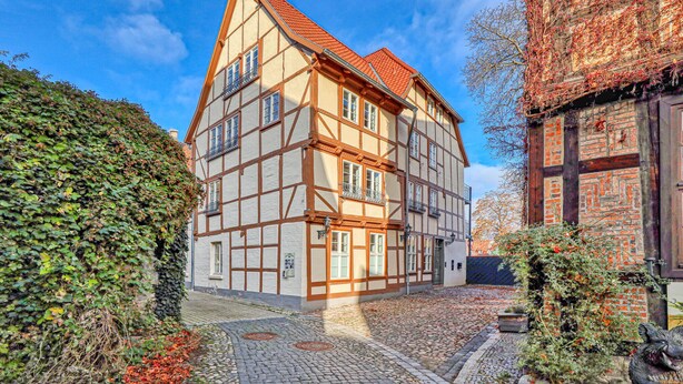 Ein traditionelles Fachwerkhäuschen mit einem steilen Satteldach, einem Kopfsteinpflasterhof und einem Baum mit roten Blättern.