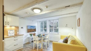 Ein gemütliches Zimmer mit Holzboden, einem gelben Sofa, einem Esstisch mit Stühlen, einer Küchenzeile mit weißen Schränken und einem großen Fenster mit Blick auf Grünflächen.
