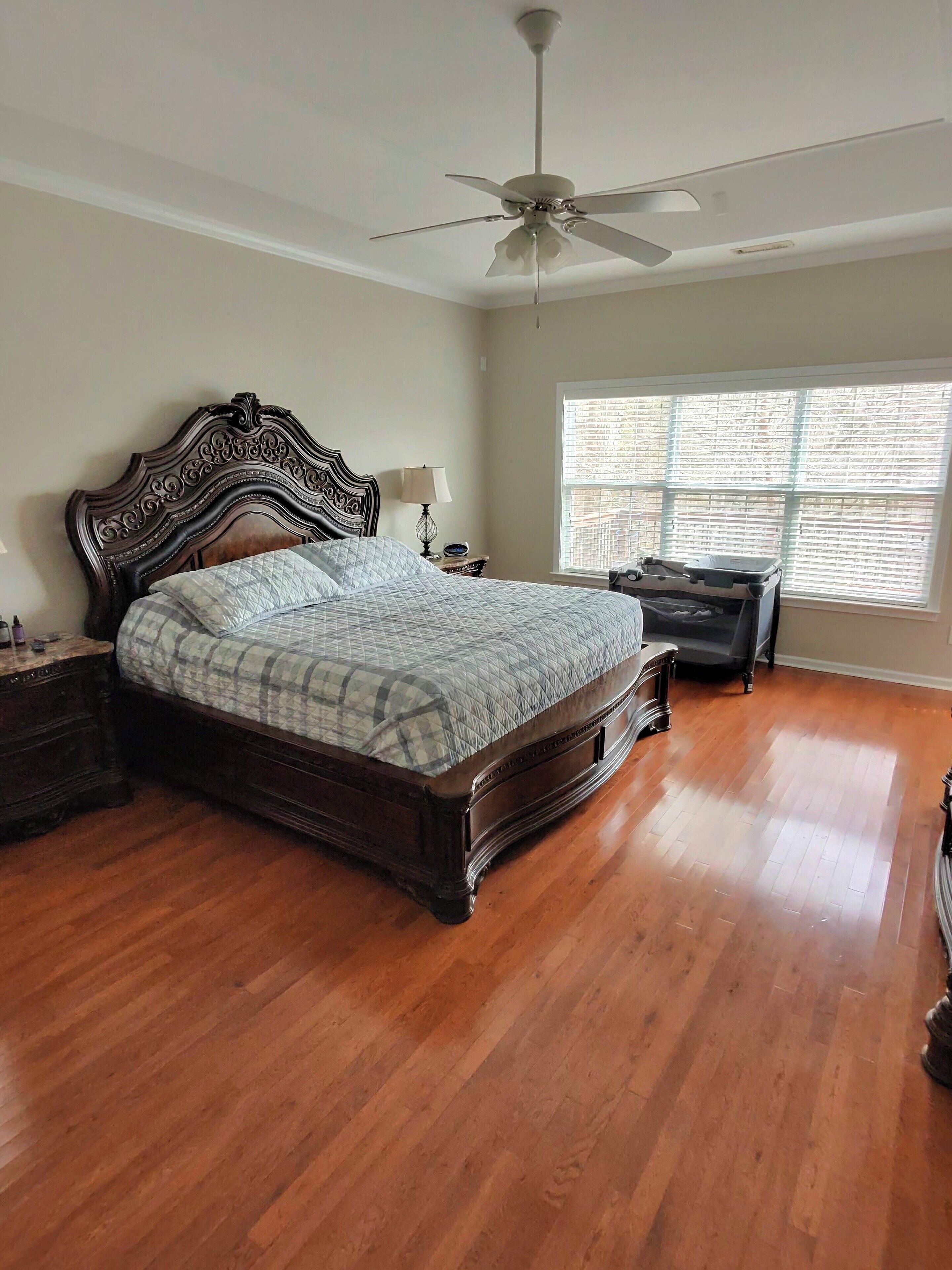 A bedroom with a large bed, a ceiling fan, a nightstand with a lamp, a dresser, and a window with blinds.