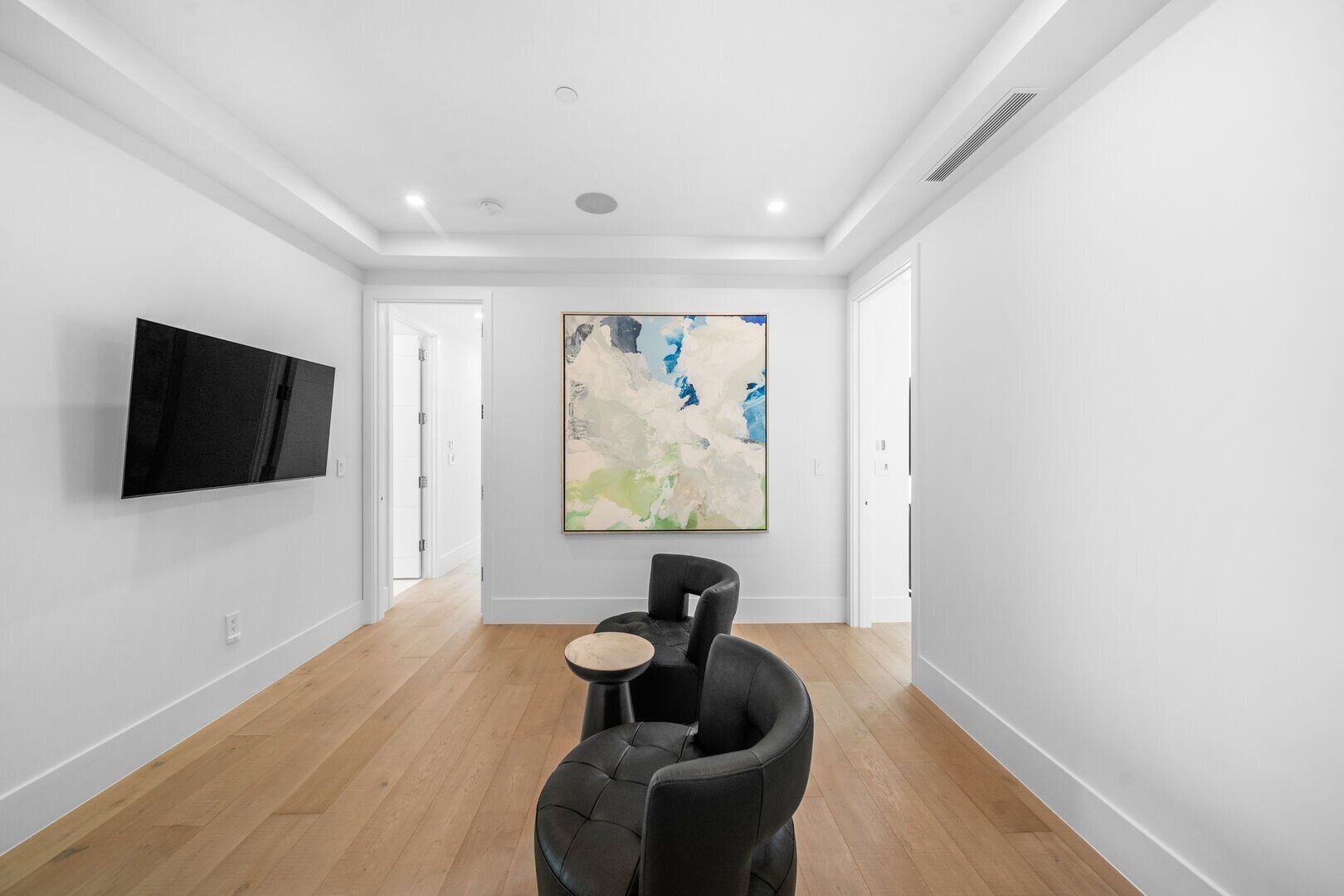 A spacious room with a flat-screen TV mounted on the wall, a large abstract painting, a black leather sofa, and a small round table.