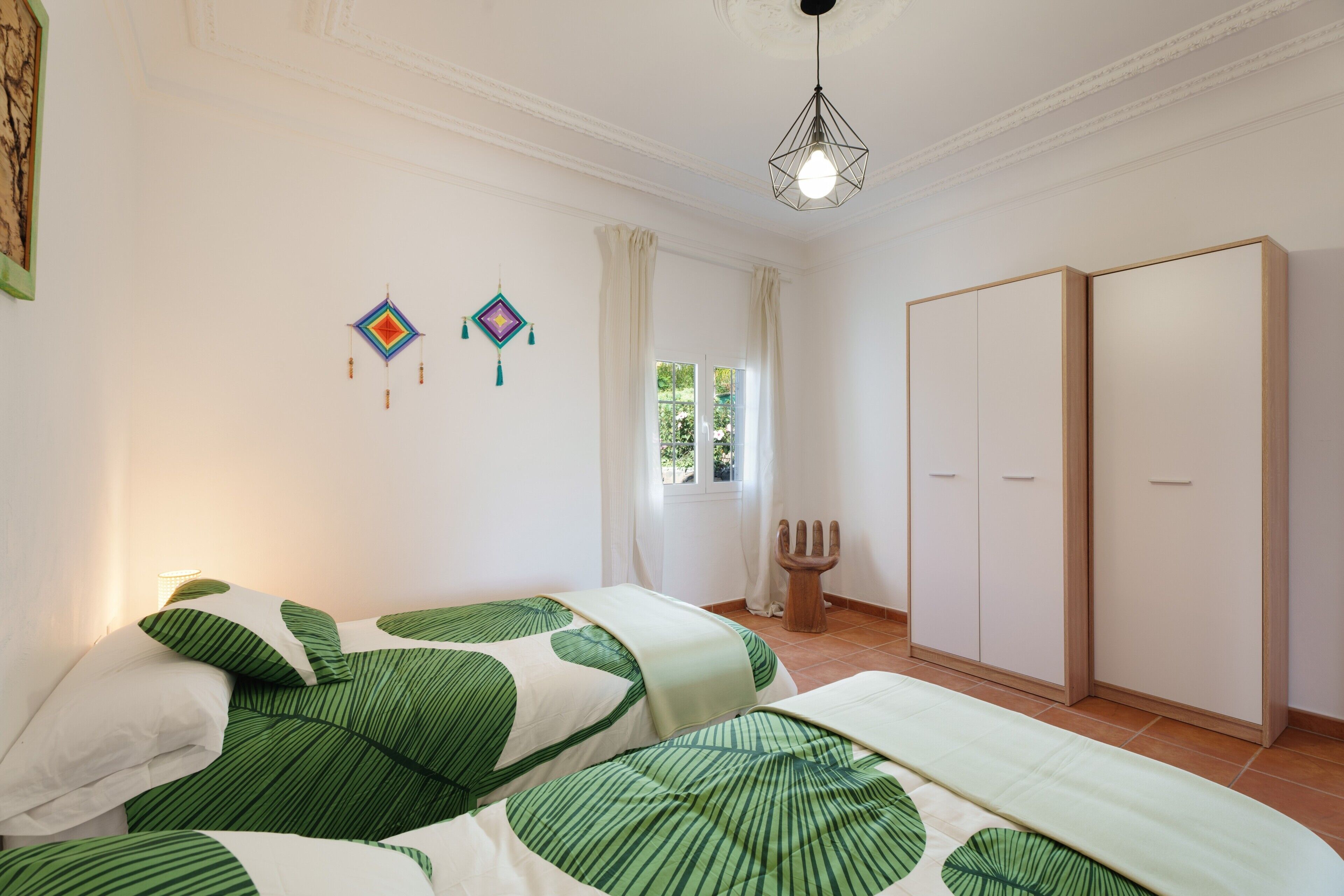 A bedroom with two beds, a wardrobe, a hanging light fixture, and a window with curtains.
