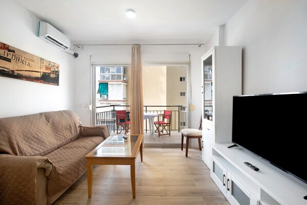 Una sala de estar con sofá, una mesa de centro de vidrio, televisor de pantalla plana y balcón con sillas rojas y blancas.