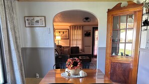 Ein Essbereich mit einem Holztisch, schwarzen Stühlen und einer floralen Tischdekoration. Eine Bank befindet sich links, im Hintergrund steht eine Lampe und eine Holztür mit Glasfeldern.