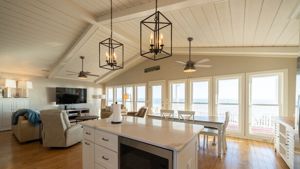 A spacious kitchen with a large island, white cabinetry, and pendant lighting. There is a dining area with a table and chairs, and a TV mounted on the wall. - 🌊 Luxe Oceanfront Escape 4BR + Private Pool & Stunning Views (Holden Beach)