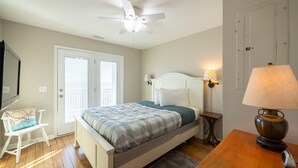 A bedroom with a bed, a ceiling fan, a nightstand with a lamp, a chair, and a door. - 🌊 Luxe Oceanfront Escape 4BR + Private Pool & Stunning Views (Holden Beach)