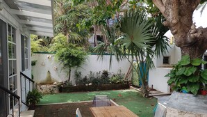 A patio with a wooden table and chairs, surrounded by lush greenery and a white wall.