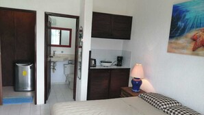 A hotel room with a bed, a desk with a lamp, a kitchenette, and a bathroom visible through an open door.