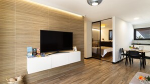 Un salon moderne doté d’un téléviseur à écran plat, d’un meuble de rangement blanc, d’un coin repas avec une table et des chaises, et d’une chambre visible à travers une porte en verre.
