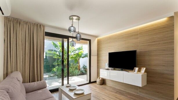 Un salon moderne avec un canapé, une table basse, un téléviseur fixé au mur en bois et une grande fenêtre avec des rideaux.